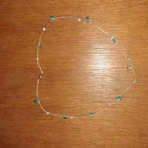 Tunt halsband med blå pärlor - Ett tunt och diskret halsband med små blå och vita pärlor på genomskinlig tråd. Pärlorna är oregelbundet placerade och ger ett minimalistiskt och fräscht intryck. Perfekt för dig som gillar enkla smycken med en touch av färg.