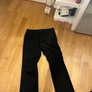 Svarta kostymbyxor med raka ben - Svarta kostymbyxor med klassisk rak passform och hällor för bälte. Byxorna har diskreta fickor fram och är sydda i ett slätt tyg som känns dressat. Perfekta för en clean och stilren look. det är smått slitet i byxan vid fötterna, byxorna är från bikbok och är lågmidjade 