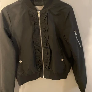 Svart bomberjacka med volangdetalj - Svart bomberjacka med dragkedja framtill och snygga volangdetaljer längs dragkedjan😊