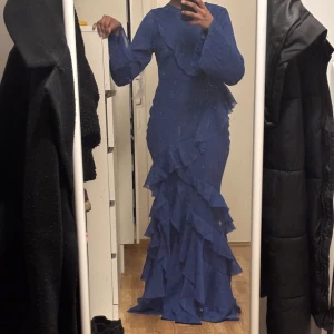 Blå långklänning med volanger - Helt ny (prislapp sitter kvar). Säljer en mörkblå långklänning med långa ärmar och massor av volanger som ger en riktigt snygg look. Klänningen har en figurnära passform och är perfekt för dig som vill sticka ut. Materialet känns lätt och flowy. 