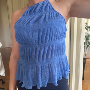 Blå plisserad linnetopp med volang - Supersöt blå topp med plisserat, liite genomskinligt tyg och volangkant nertill. Toppen har tunna axelband och en luftig, flowy passform som är perfekt nu för sommaren. Snygg halterneck-modell som ger en fräsch och trendig look. 💓💓💓💓