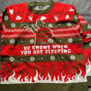Freddy Krueger jultröja XXL - Säljer en grön och röd stickad jultröja med Freddy Krueger-motiv och texten 'He knows when you are sleeping'. Tröjan har snöflingor, eldflammor och vita detaljer, samt klassiskt crewneck. Perfekt för skräckfilmsfans som vill sticka ut på julfesten.