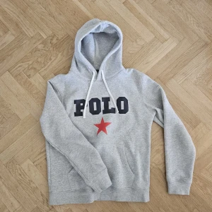 Grå hoodie från Polo med stjärna - Snygg ljusgrå hoodie från Polo Ralph Luaren med stor mörkblå logga och röd stjärna på bröstet. Tröjan har huva med vita snören. Använd fåtal gånger och har inga defekter. Skriv vid frågor eller funderingar. Kan skicka fler bilder om man vill ha det. ☺️🔥