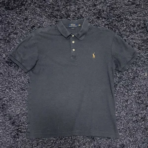 Mörkgrå pikétröja Polo Ralph Lauren - Snygg mörkgrå pikétröja från Polo Ralph Lauren i slim fit. Klassisk design med krage, tre knappar och den ikoniska broderade polospelaren på bröstet. Perfekt för en clean och stilren look. Tillverkad i mjuk bomull för skön känsla.