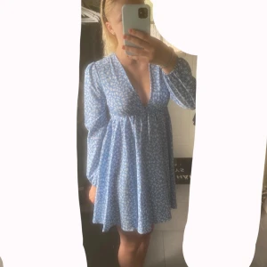 Blå blommig kortklänning med v-ringning - Supersöt blå kortklänning med vita blommor, djup v-ringning och långa ärmar. Klänningen har en flowy passform och dragkedja bak. Perfekt för sommarens alla tillfällen.