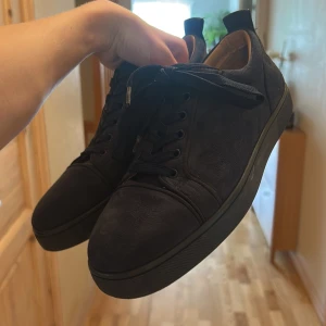 Mörkblå sneakers från Christian Louboutin - Snygga mörkblå sneakers från Christian Louboutin i mocka med klassisk röd sula. Skorna har rund tå, platt sula och svarta snören. Insidan är ljusbeige och de har diskreta detaljer som gör dem riktigt stilrena. Perfekta för dig som vill sticka ut med lyxig känsla.