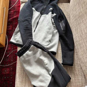 Nike Tech Fleece zip hoodie svart/grå - Nike Tech Fleece hoodie i svart och ljusgrått med hel dragkedja och huva. Snygg kontrast mellan färgerna, vit Nike-logga på bröstet och praktisk ficka med dragkedja på ärmen, riktigt skön och modern passform. Tröjan är storlek S och byxorna storlek M 