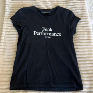 Svart Peak Performance t-shirt XS - Svart t-shirt från Peak Performance med vit logga och text 'Est. 1986' på bröstet. Klassisk rund halsringning och korta ärmar. Skön och enkel modell i mjuk bomull, perfekt till jeans eller shorts.