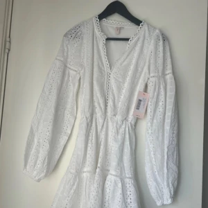 Vit spetsklänning med lång ärm - Superfin vit kortklänning från Nelly med broderade spetsdetaljer och v-ringning. Klänningen har långa ärmar med resår vid handlederna och en luftig passform. Perfekt för sommarens alla tillfällen!