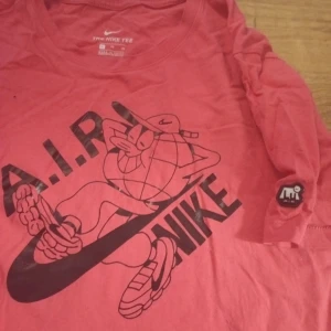 Nike AIR x Lugosis röd långärmad tee XL - Nike AIR Artist in Residence tee designad av Lugosis i rött med svart tryck framtill. Unik illustration med globfigur och Nike-logga, samt AIR-märke på ärmen. Långärmad modell i mjuk bomull, perfekt för dig som gillar streetwear och coola prints.