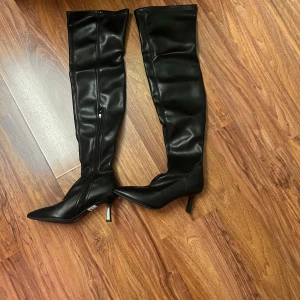 Svarta overknee stövlar med klack - Snygga svarta overknee stövlar i skinnimitation med spetsig tå och smal klack. Dragkedja på insidan för enkel påtagning. Perfekta för att lyfta din outfit med en edgy vibe. Passar dig som gillar statement boots och stilren look.