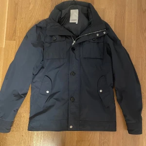  J.Lindeberg Jacka  - Snygg mörkblå vindjacka från J.Lindeberg med klassisk krage, flera fickor med knappar och dragkedja. Jackan har även en luva i kragen du kan ta ut eller ha kvar. Skriv för mer bilder osv!