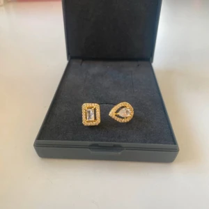 Guldig ring med stenar - Unik guldig ring med två olika former framtill – en rektangulär och en droppformad detalj, båda kantade av små glittrande stenar. Ringen har öppen design och ger en lyxig känsla med sina gnistrande detaljer. Perfekt för dig som vill sticka ut med accessoarer.