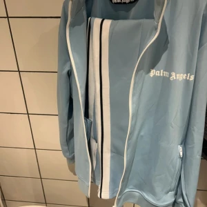 Blå Palm Angels track dress - Snygg ljusblå track jacket från Palm Angels med vita och svarta ränder längs ärmarna. Jackan har dragkedja framtill, fickor med dragkedja och vit logga på bröstet. Perfekt för en sportig och trendig look. Byxorna är det M på. Tröjan är i S. Helt ny. Hade tyvärr beställt fel storlek på byxor…