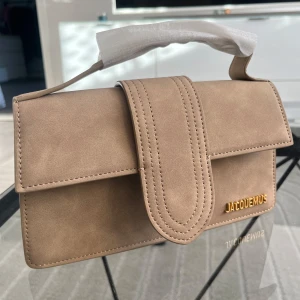 Beige handväska från Jacquemus - Snygg beige handväska från Jacquemus med stilren design och guldfärgad logga framtill. Väskan har ett brett lock med dekorativa sömmar, praktiskt handtag och en ficka på baksidan. Tillverkad i mjuk mocka som ger en lyxig känsla. Väskan är inte hos mig så kan ej skicka mer bilder än detta. Priset kan ej diskuteras. Nej finns inget kvitto. 