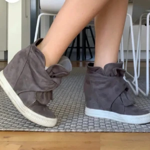 Gråa kilklack skor - Snygga grå sneakers med kilklack och stor rosettdetalj framtill. Skorna har vit platt sula och är gjorda i mockaliknande material. Perfekta för dig som vill ha en cool och unik look med extra höjd.