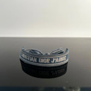 Blått armband  - Snyggt blått armband från Christian Dior med texten 'J'Adior' broderad i vitt. Armbandet har en vävd struktur och avslutas med tofsar för en cool och avslappnad look. Perfekt accessoar för att lyfta din outfit.
