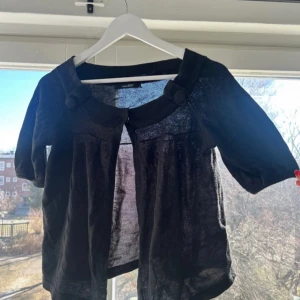 Svart bolerotopp från Vero Moda - Snygg svart bolerotopp från Vero Moda med trekvartsärm och dekorativa knappar vid halsringningen. Perfekt för att lägga till en stilfull touch till din outfit. Tillverkad i ett lätt och luftigt material.