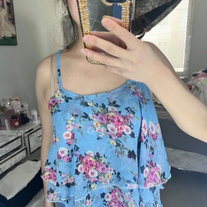Blommig blå sommar topp - Säljer en söt blå klänning med blommigt mönster i rosa och lila. Klänningen har smala axelband och volangdetaljer som ger en romantisk känsla. Perfekt för varma sommardagar!