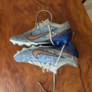 Nike mercurial cr7 - Snygga fotbollsskor från Nike med ett unikt blått och vitt mönster. Skorna har snörning och en distinkt Nike-logga i orange. Perfekta för fotbollsplanen med sina greppvänliga dobbar.