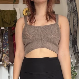 Brun stickad croppad topp - Köpt secondhand men aldrig använt. Säljer en brun stickad croppad topp med breda axelband. Toppen har en unik design med en öppen del framtill som ger en modern look. Perfekt för att styla med höga byxor eller kjolar.