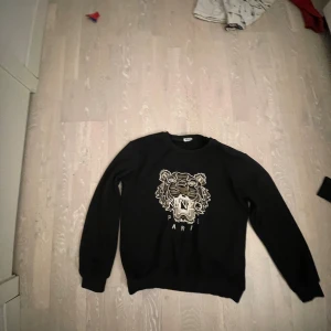 Svart tröja från Kenzo med tigerbroderi - Säljer en svart tröja från Kenzo med ett stort broderat tigerhuvud på framsidan. Tröjan har långa ärmar och en rund halsringning. Perfekt för en stilren och trendig look.