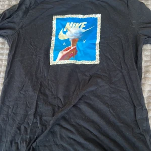 Svart t-shirt med tryck från Nike - Snygg svart t-shirt från Nike med ett färgglatt tryck av en glass och texten 'Nike Air'. Perfekt för en avslappnad stil. T-shirten har korta ärmar och är gjord i ett bekvämt material.