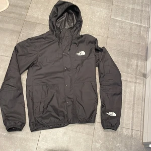 Svart vindjacka från The North Face - Säljer en svart vindjacka från The North Face med huva och dragkedja. Jackan har långa ärmar och tryckknappar framtill. Perfekt för blåsiga dagar. Passar upp till S