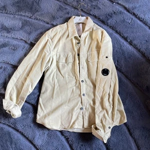 Beige manchester skjorta från C.P. Company - Snygg beige skjorta i manchester från C.P. Company. Skjortan har långa ärmar och knappar framtill samt två bröstfickor. Perfekt för en stilren look. 2 mindre fläckar på ryggen som du kan se på bild 3 men annars är den varsamt använd få gånger och är perfekt nu inför sommaren! Hör av er ifall ni har frågor😇