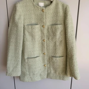 Ljusgrön kavaj med guldknappar bouclé - Elegant ljusgrön kavaj i ett texturerat tyg med stiliga guldknappar framtill. Kavajen har en klassisk design med fyra fickor och rund halsringning. Perfekt för att ge en sofistikerad touch till din outfit. Storlek S från H&M