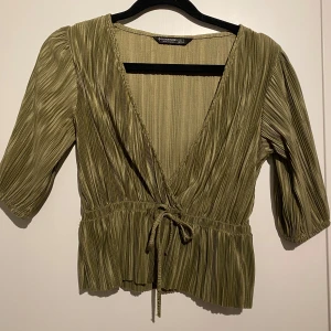 Olivgrön omlottopp från Stradivarius! S - Säljer en olivgrön omlottopp  med trekvartsärmar och knytband i midjan. Toppen har en v-ringad design och ett plisserat mönster som ger en elegant touch. Perfekt för en stilren look. Superfint skick! 