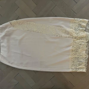 Vit kjol med spetsdetaljer - Elegant vit kjol med vackra spetsdetaljer längs sidorna. Kjolen har en mjuk och flytande passform som ger en feminin touch. Perfekt för en stilren look. Inköpt 2024 och använd 1 ggr. Cremevit färg