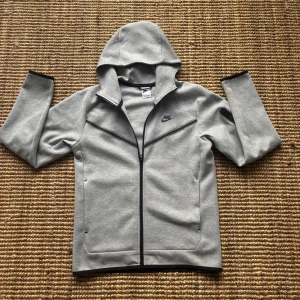 Nike tech ny (m kvitto) - Säljer en stilren Nike tech hoodie med dragkedja och huva. Jackan har långa ärmar och en diskret Nike-logga på bröstet. Perfekt för en sportig look eller avslappnade dagar. Helt nyskick och använd 1 gång. Pris ej hugget i sten.