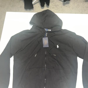 Svart hoodie från Polo Ralph Lauren - Säljer en stilren svart hoodie från Polo Ralph Lauren med dragkedja och det klassiska logotypbroderiet på bröstet. Perfekt för en avslappnad look. Hoodien har långa ärmar och en bekväm passform. Den kan också passa S