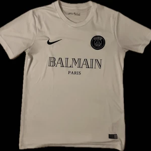 Vit t-shirt från Balmain Paris för 200kr - Snygg vit t-shirt från Balmain Paris med korta ärmar och rund hals. T-shirten har en stilren design med Balmain Paris-logga och Nike-märke på framsidan. Perfekt för en casual look.