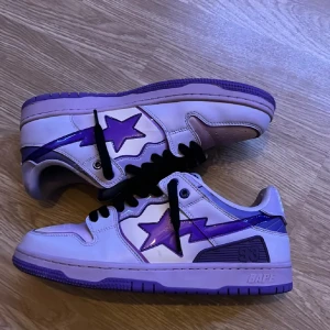 Lila och vita sneakers från Bape - Snygga Bape sneakers i en cool kombination av lila och vitt. Skorna har en stjärndetalj på sidan och en robust sula med stjärnmönster. 