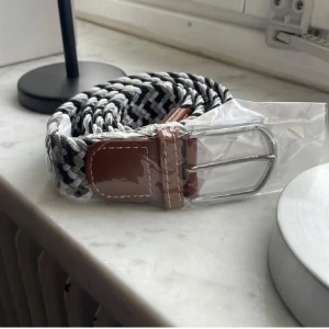 Flätat bälte  - Snyggt flätat bälte i svart, grått och vitt med bruna läderdetaljer och ett silverfärgat spänne. Perfekt för att ge en stilren touch till din outfit. 105 cm långt och passar alla då det är väldigt elastiskt.