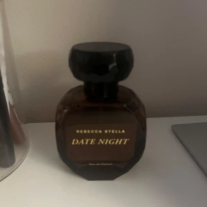 Date Night Eau de Parfum från Rebecca Stella - Elegant parfymflaska i mörkbrunt glas med en facetterad design och svart lock. Doften 'Date Night' ger en lyxig och förförisk känsla, perfekt för speciella tillfällen. Flaskan har en stilren etikett med guldtext som ger en exklusiv touch.