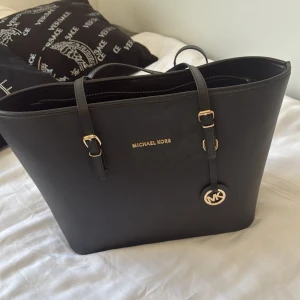 Svart axelväska från Michael Kors - Snygg svart axelväska från Michael Kors med gulddetaljer och logotyp. Väskan har dubbla handtag och en praktisk dragkedja för säker förvaring. Perfekt för att bära med sig det nödvändigaste med stil. Äkta väska, säljer den billig då den inte har användts på länge