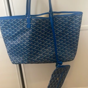 Blå mönstrad shoppingväska - Snygg blå shoppingväska med ett ikoniskt mönster och matchande liten plånbok. Väskan har långa handtag och en öppen design med ljus insida. Perfekt för att bära med sig allt man behöver under dagen.