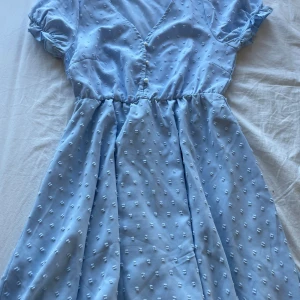 Ljusblå klänning med knappar - Charmig ljusblå klänning med korta ärmar och v-ringning. Klänningen har ett diskret prickigt mönster och knappar framtill. Perfekt för sommarens varma dagar.