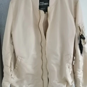 Beige bomberjacka från Alpha Industries - Snygg beige bomberjacka från Alpha Industries med dragkedja framtill och fickor med knappar. Jackan har en klassisk bomberstil med långärmade ärmar och en liten ficka med dragkedja på ärmen. Perfekt för en stilren look.