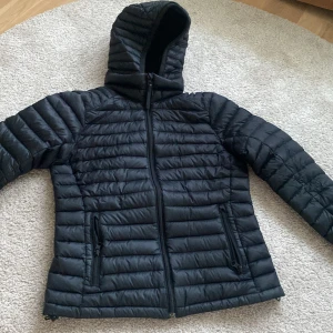 Svart dunjacka från Peak Performance - Säljer en jättefin svart dunjacka från Peak Performance med huva och dragkedja. Jackan har en quiltad design och två praktiska fickor med dragkedjor. Perfekt för kyliga dagar! Avtagbar luva tillkommer! Finns ett litet hål på armen men det syns knappt när man har på sig jackan!!