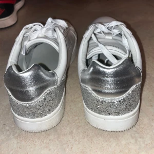 Vita sneakers från ONLY med silverdetaljer - Snygga vita sneakers från ONLY med glittriga silverdetaljer på hälen. Skorna har snörning och en klassisk design som passar perfekt för en trendig look. Perfekta för dig som vill ha en stilren men ändå iögonfallande sko. Endast använda 1 gång.💕
