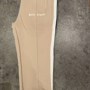 Beige byxor från Palm Angels - Snygga beige byxor från Palm Angels med vit sidoremsa. Byxorna har en modern och stilren design med en avslappnad passform. Perfekta för en trendig look.