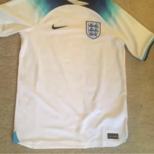 Engelsk fotbollströja - Säljer en vit fotbollströja från Nike med Englands landslagsemblem på bröstet. Tröjan har korta ärmar och ett snyggt blått mönster vid axlarna. Perfekt för fotbollsfans som vill visa sitt stöd för laget! ⚽