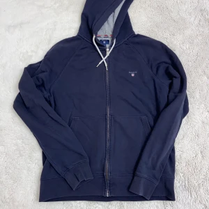 Mörkblå hoodie från Gant passar även L och M med något ovanpå. - Säljer en snygg mörkblå hoodie från Gant med dragkedja och justerbar huva. Tröjan har långa ärmar och en praktisk känguruficka framtill. Perfekt för en avslappnad stil.