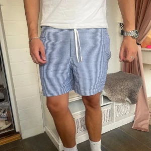 Randiga Badbyxor - Snygga blå och vita randiga badbyxor från Bondelid med snörning i midjan. De har en avslappnad passform och är tillverkade i ett lätt och bekvämt material.