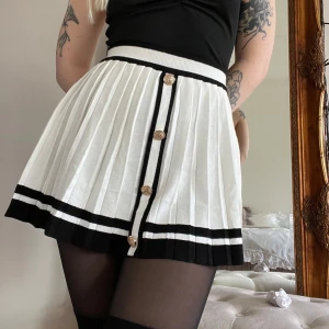 Vit och svart plisserad kjol med knappar - Snygg vit plisserad kjol med svarta ränder och dekorativa knappar framtill. Kjolen har en elastisk midja och är perfekt för en stilren look. Den är kort och har en klassisk design som passar till många olika tillfällen.