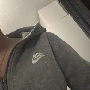 Grå hoodie från Nike - Säljer en klassisk grå hoodie från Nike med dragkedja och huva. Perfekt för en avslappnad stil. Den har en broderad Nike-logga på bröstet och är tillverkad i mjukt material för extra komfort.